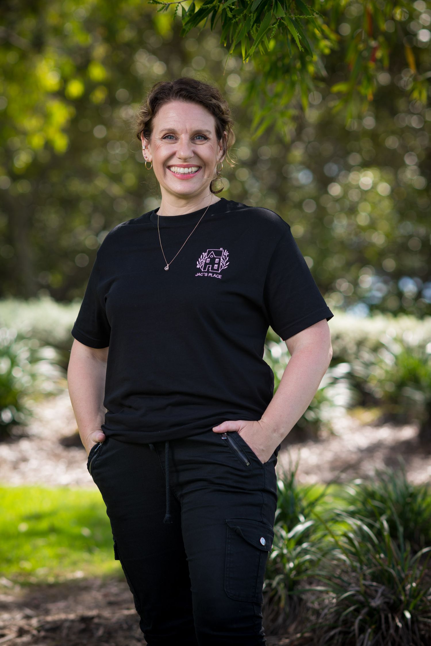 Smiling woman in black uniform stands outdoors in a garden setting with lush green trees in background.