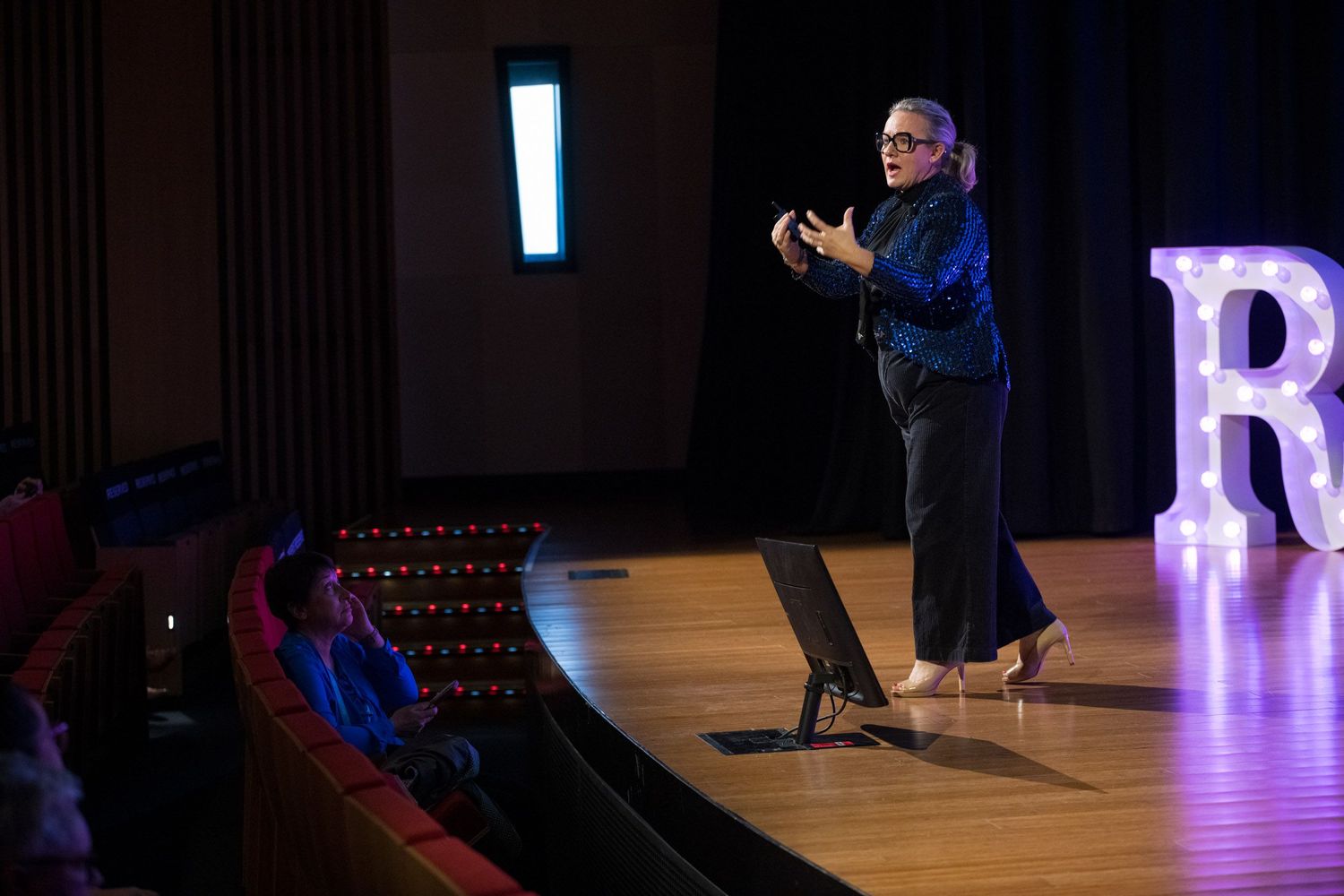 Female speaker presents to seated audience on theater stage beside illuminated letter R display.