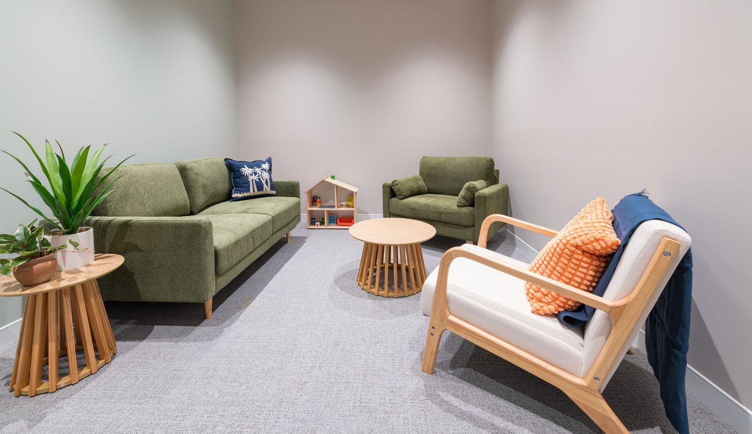 Contemporary waiting room with green velvet sofa, wooden armchairs, orange cushions, and indoor plants on grey carpet.
