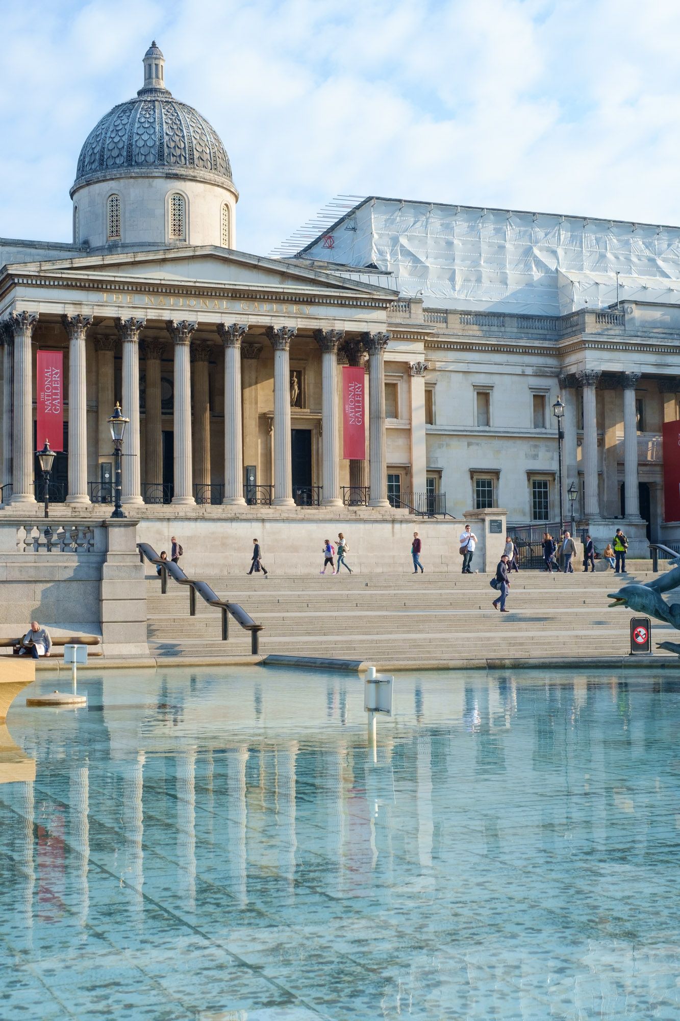 The National Gallery at Trafalgar Square in London, reflected in a fountain pool with visitors on steps.