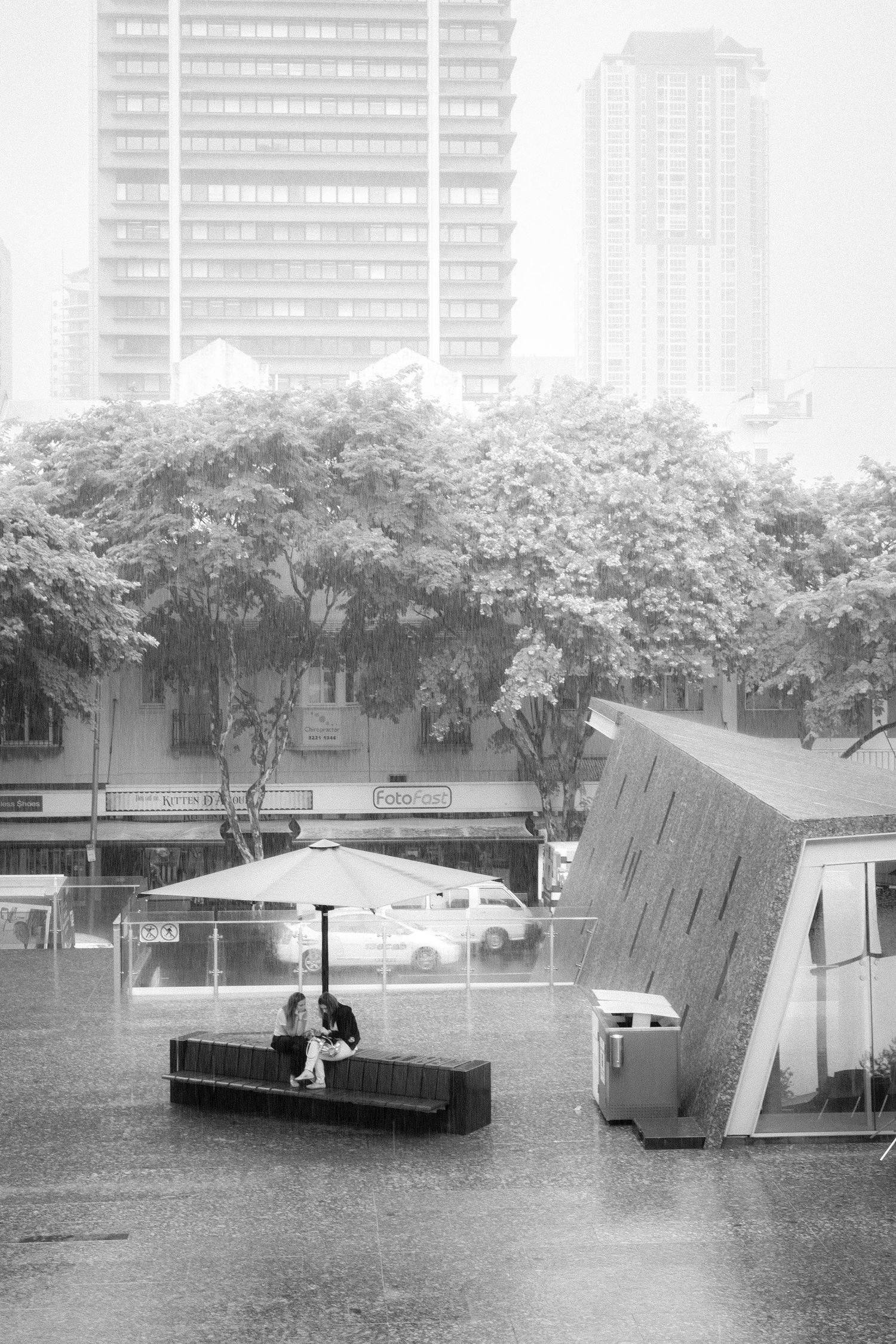 Black and white photo of a person sitting alone in heavy rain in an urban city square.