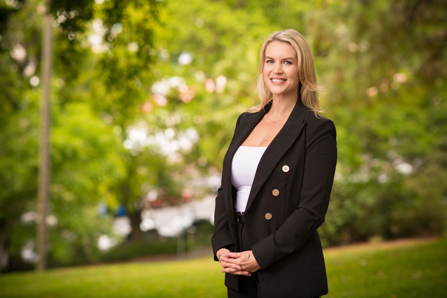 Professional blonde woman in black blazer standing outdoors in a park with green trees behind her.