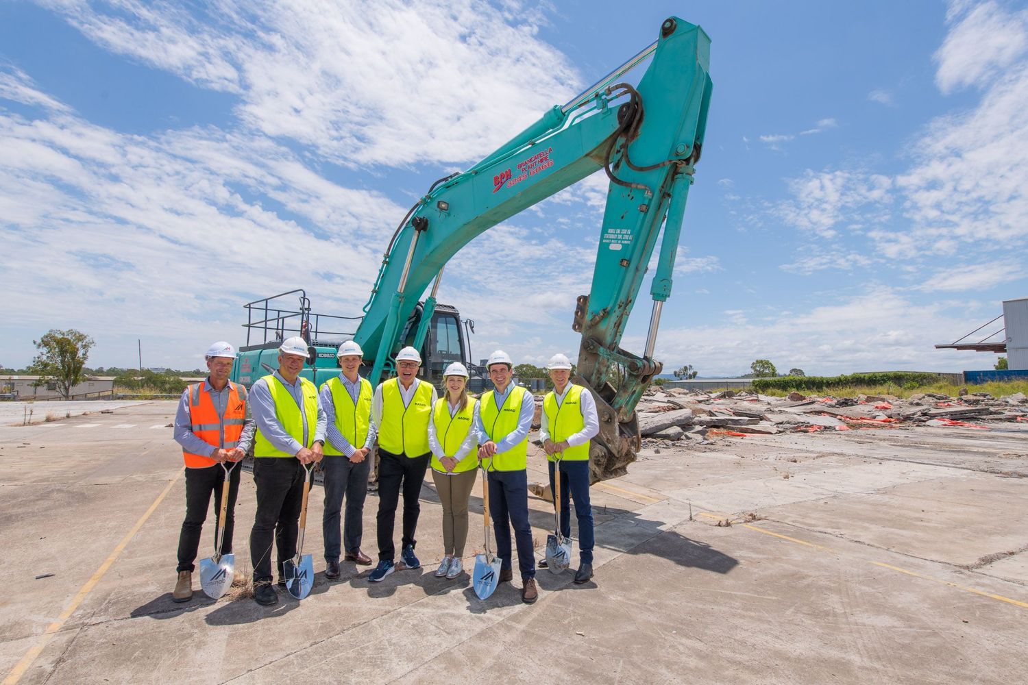 Group of six people in hi-vis vests and hard hats at construction groundbreaking ceremony with excavator.