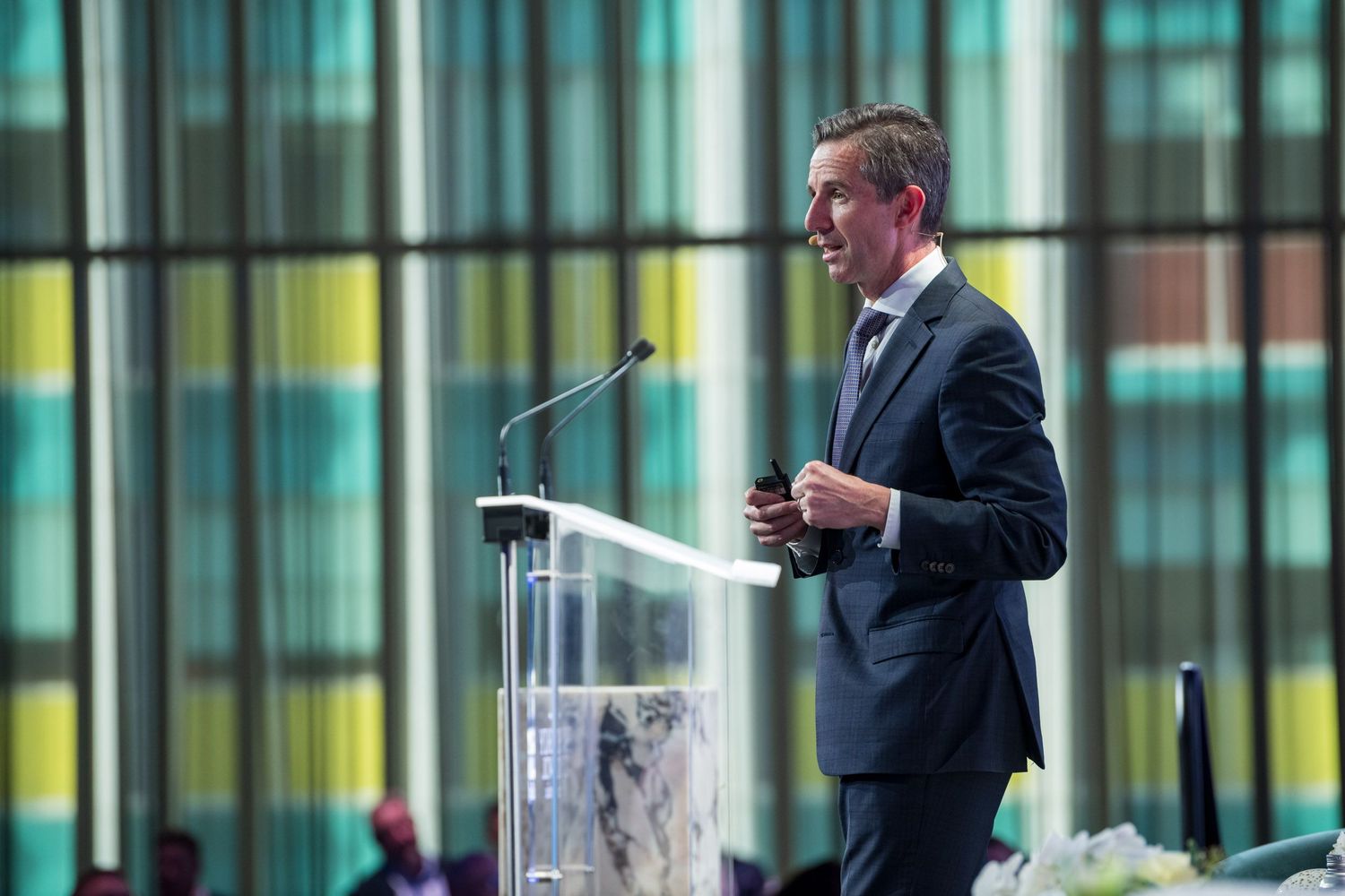 Professional male speaker addresses audience from glass podium at formal conference event.