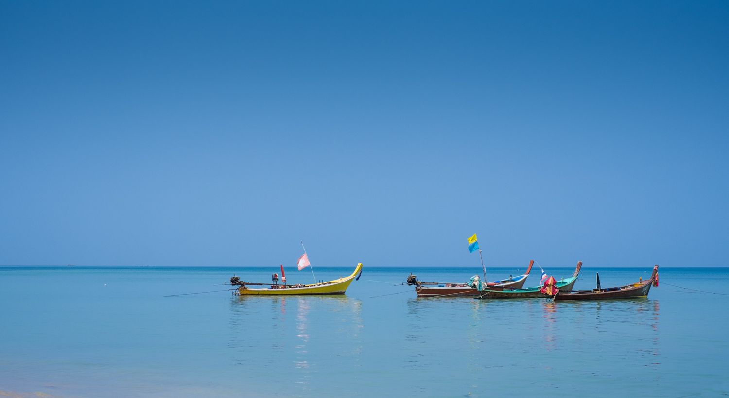 Traditional colorful longtail boats float on crystal clear turquoise water under a vivid blue sky in Thailand.