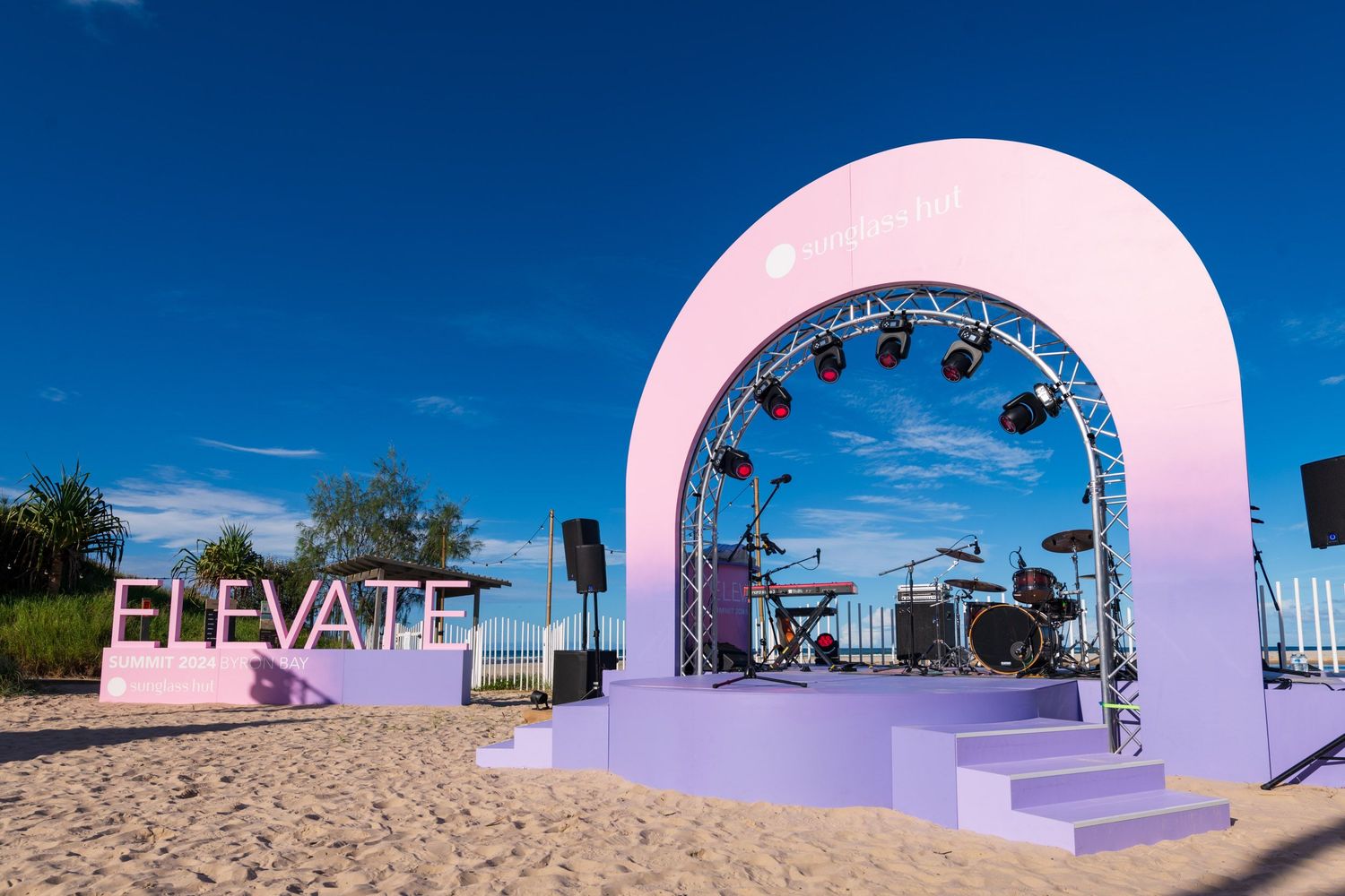 Pink arch stage with ELEVATE sign and live band setup on a sunny beach.