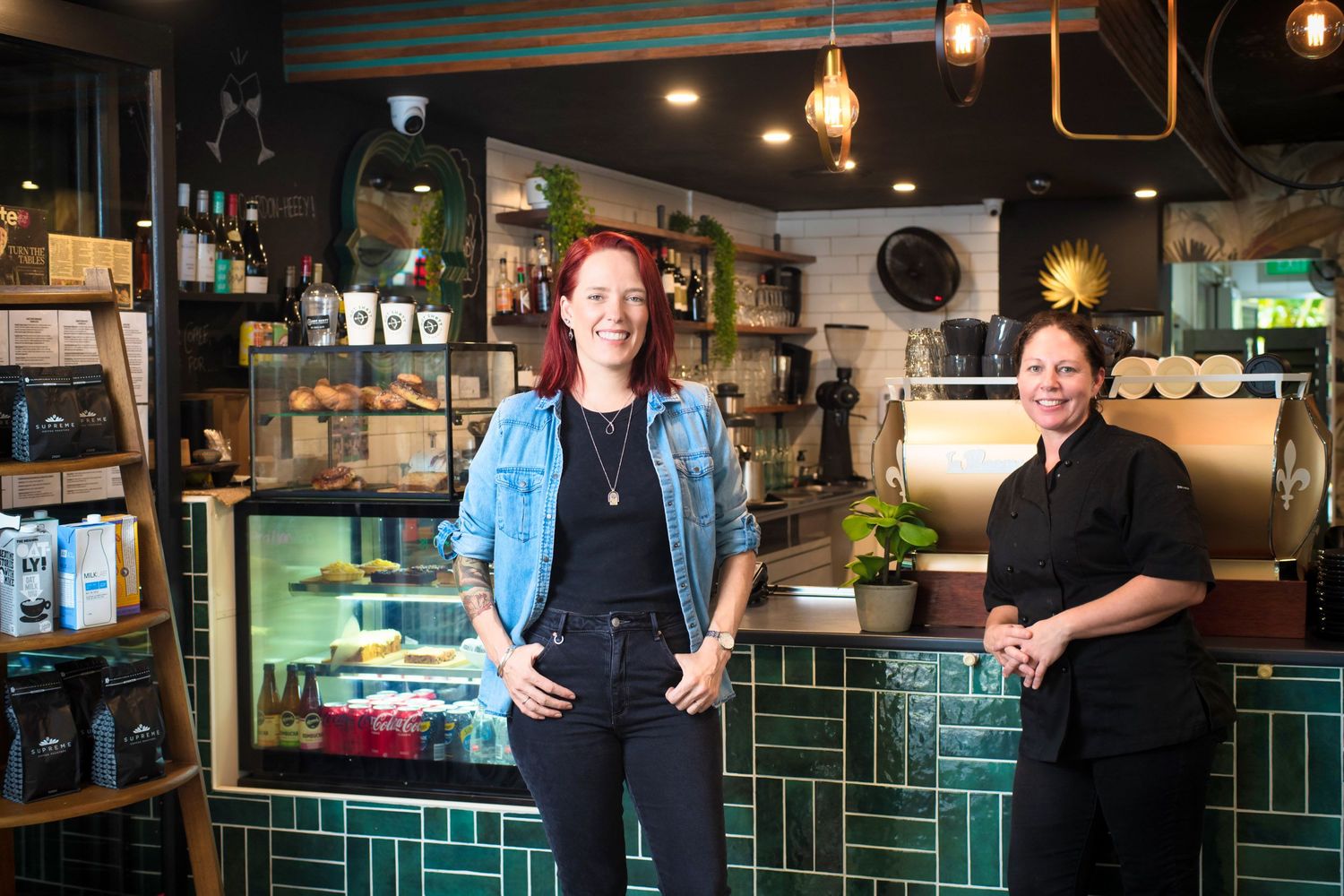 Two women smiling in trendy cafe with green tiled walls and coffee bar.