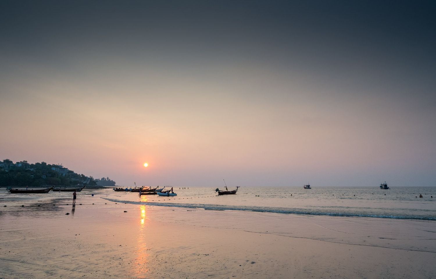 Silhouetted longtail boats rest on a tranquil beach at a hazy pastel sunset in tropical Thailand.