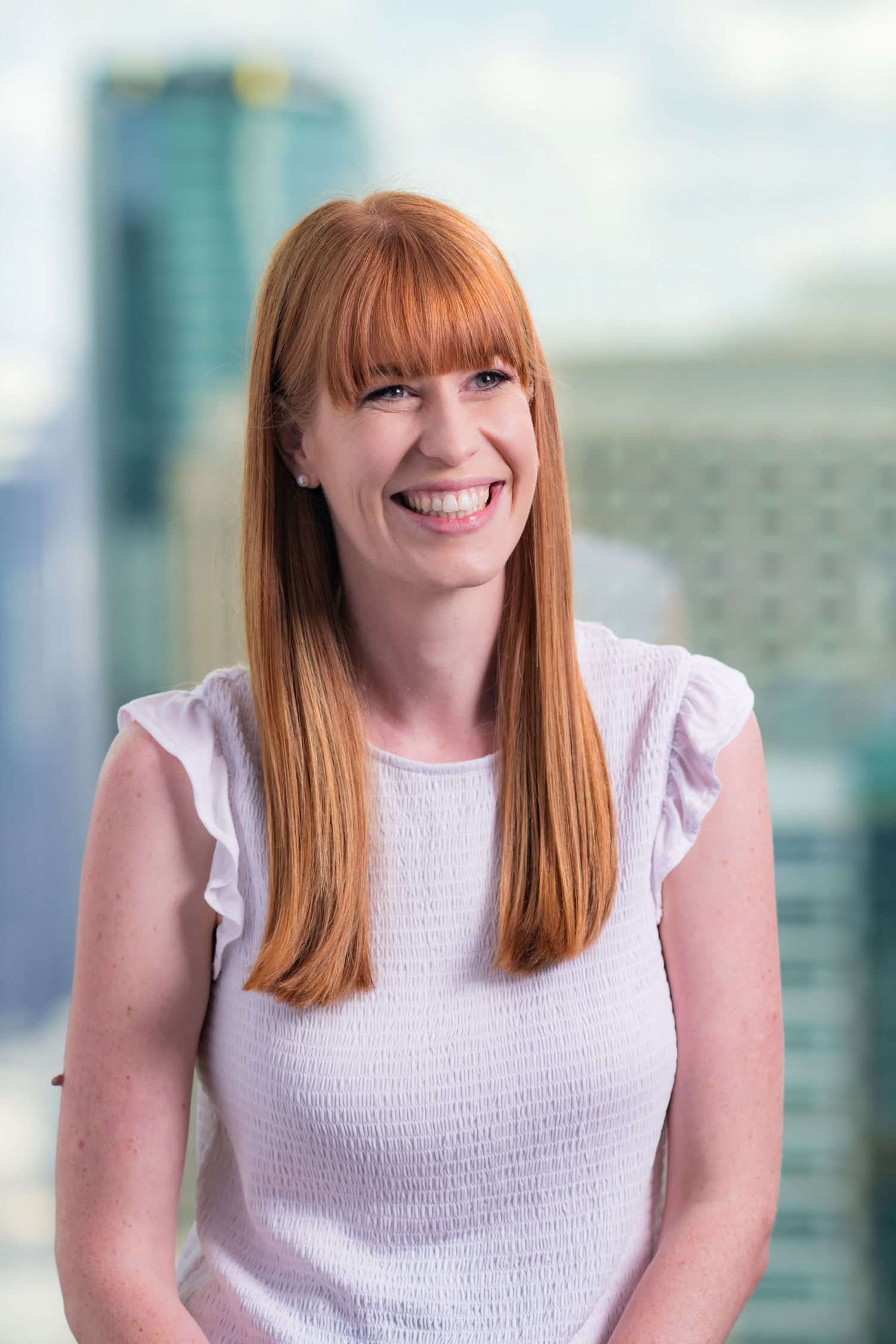 Smiling redhead woman in white top with city skyline blurred in the background behind her.