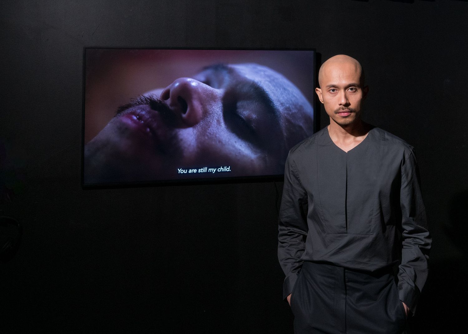 Bald man in grey outfit standing before a dark video art installation displaying emotional scene.