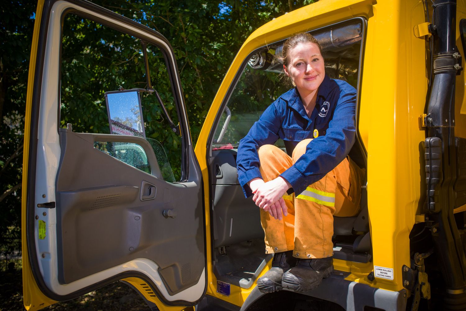 Female worker in hi-vis uniform sits in open door of yellow truck cab outdoors.