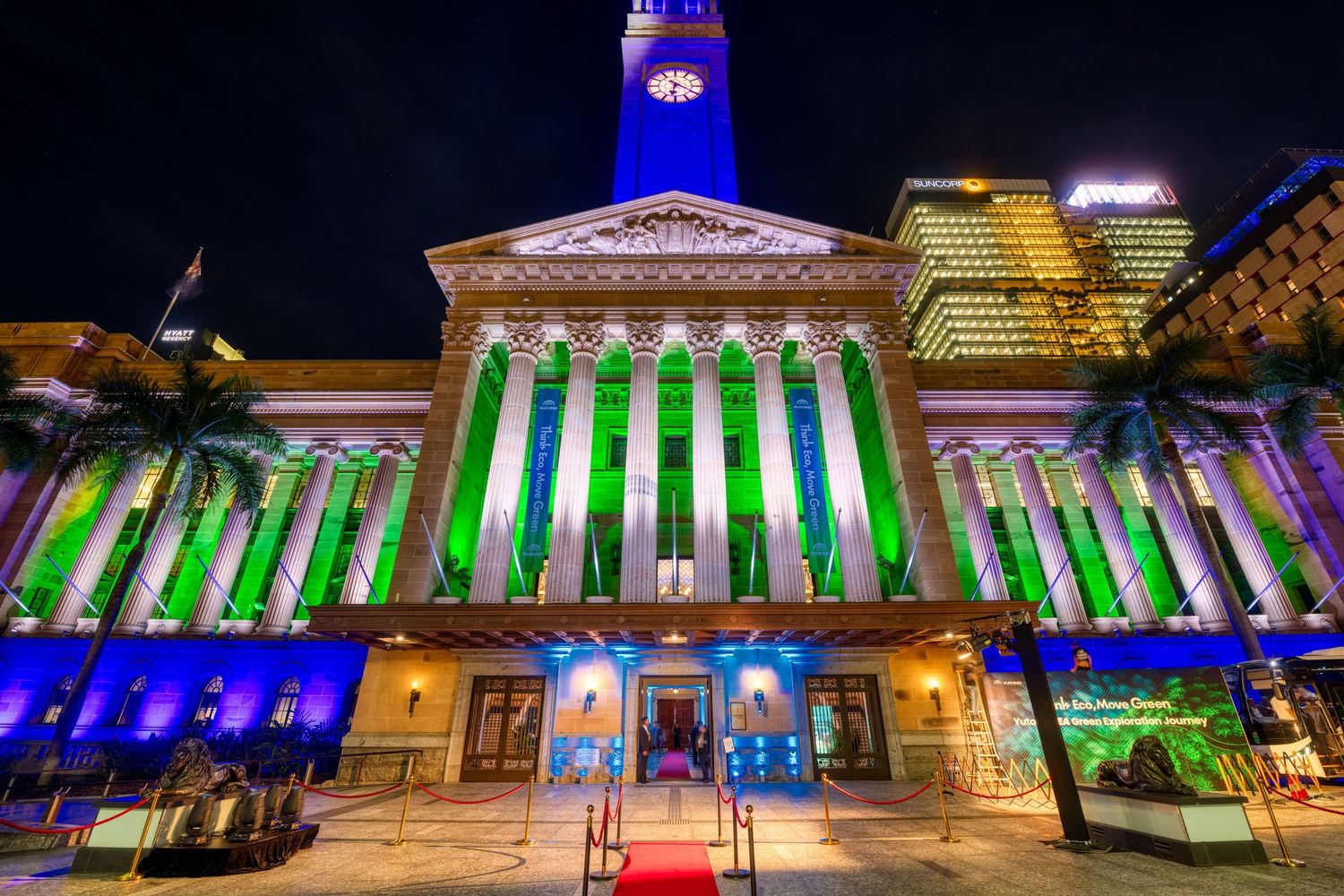 Brisbane City Hall illuminated with vibrant green and blue lights at night with palm trees and red carpet entrance.
