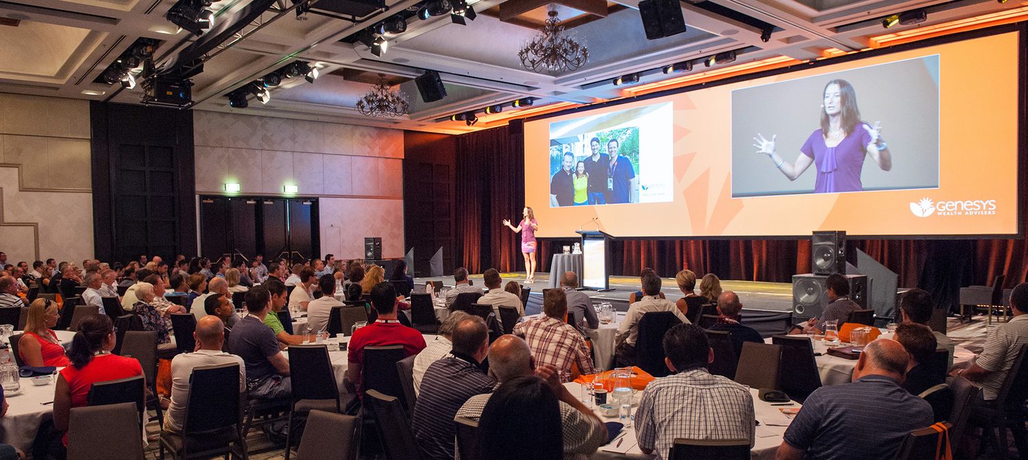 Large audience attends a professional conference presentation in a modern hotel ballroom with large screens.