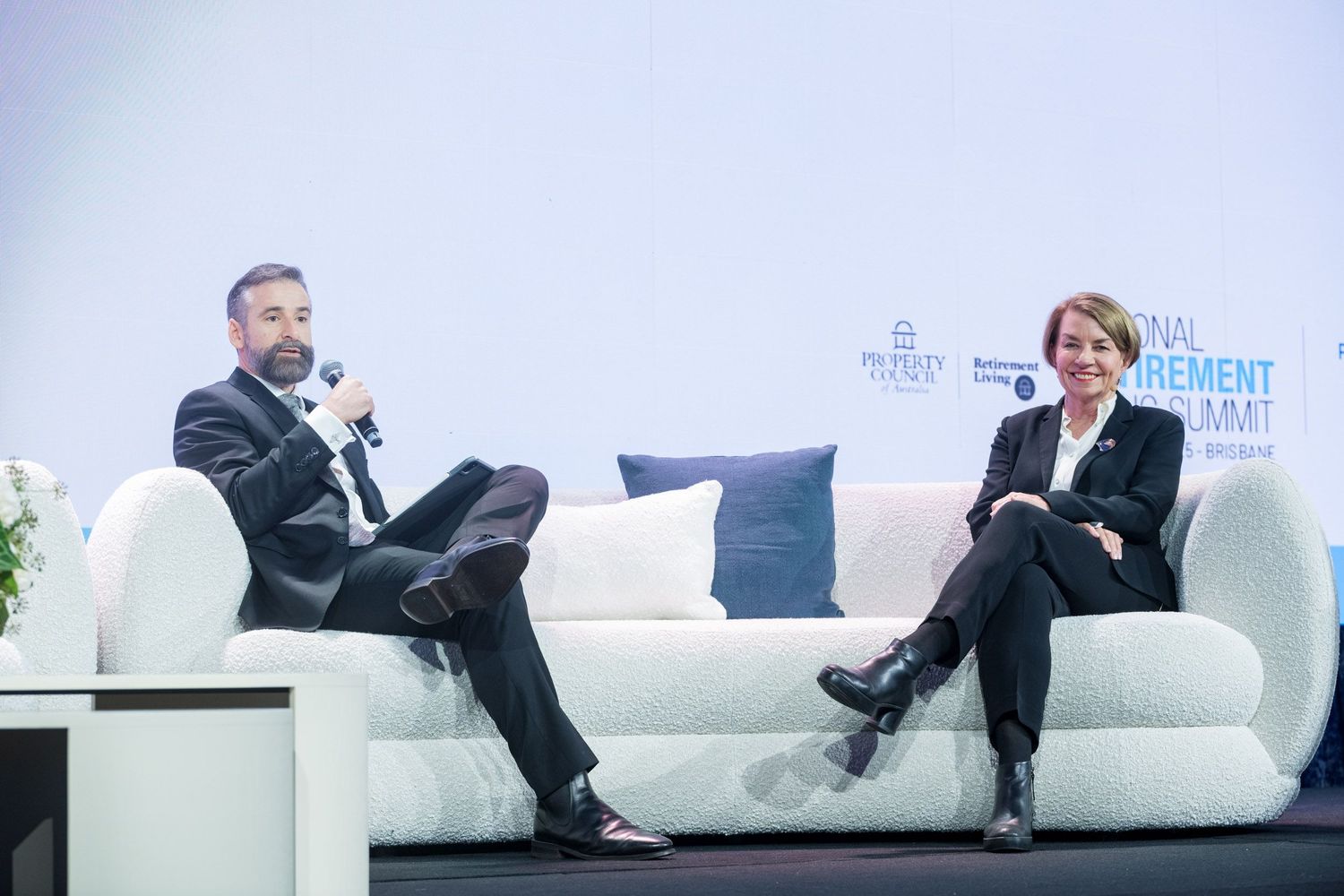 Two professionals seated on white sofa during panel discussion at management summit conference.