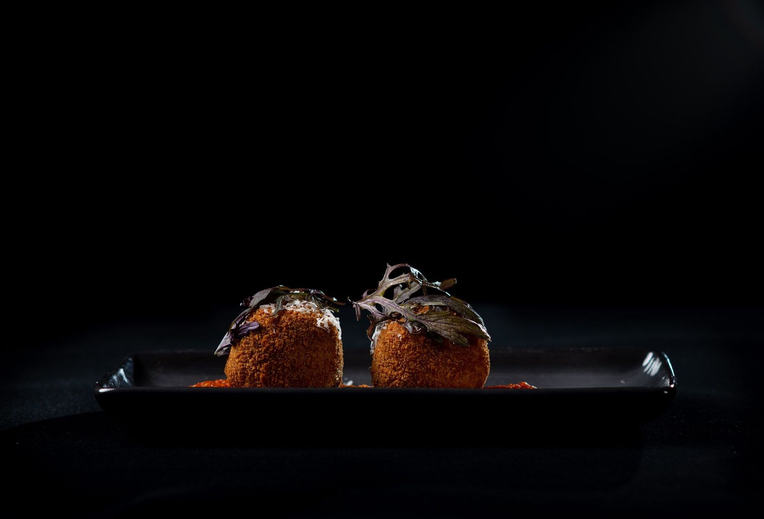 Two chocolate arancini balls dusted with cocoa powder on a dark serving plate.