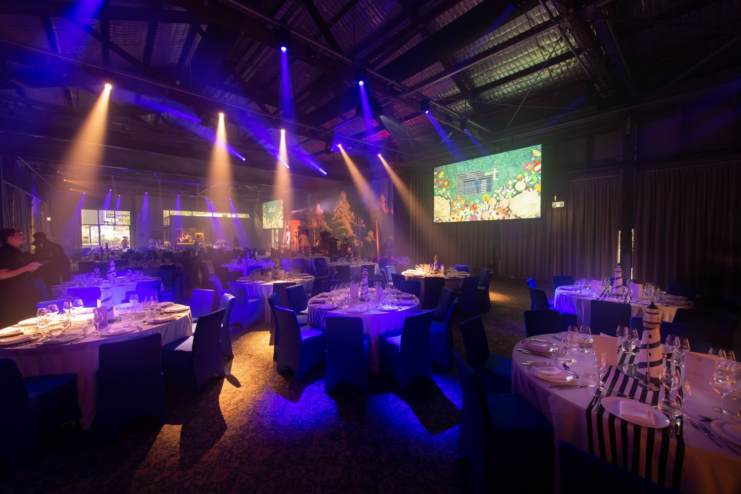 Dramatic event venue with blue uplighting, spotlights, round dining tables, and large projection screen display.
