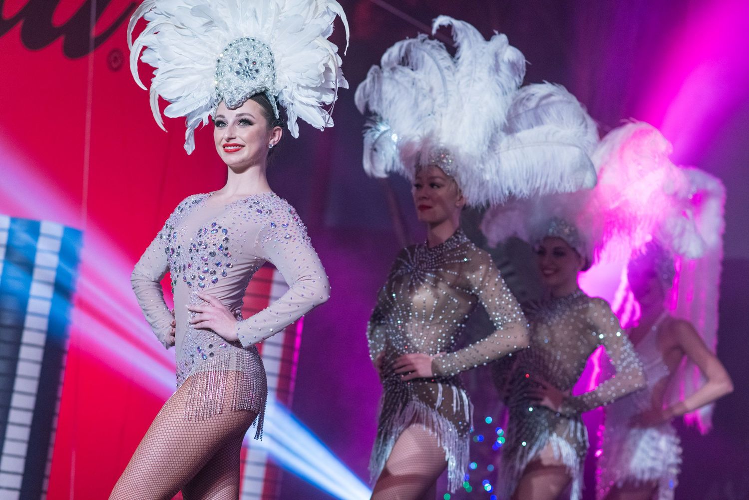 Showgirls in sparkling white costumes and feathered headdresses performing on a colorful stage.