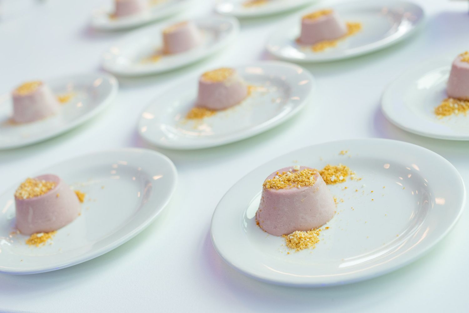 Rows of elegant plated lavender panna cotta desserts with gold crumble on white oval plates at a gala.