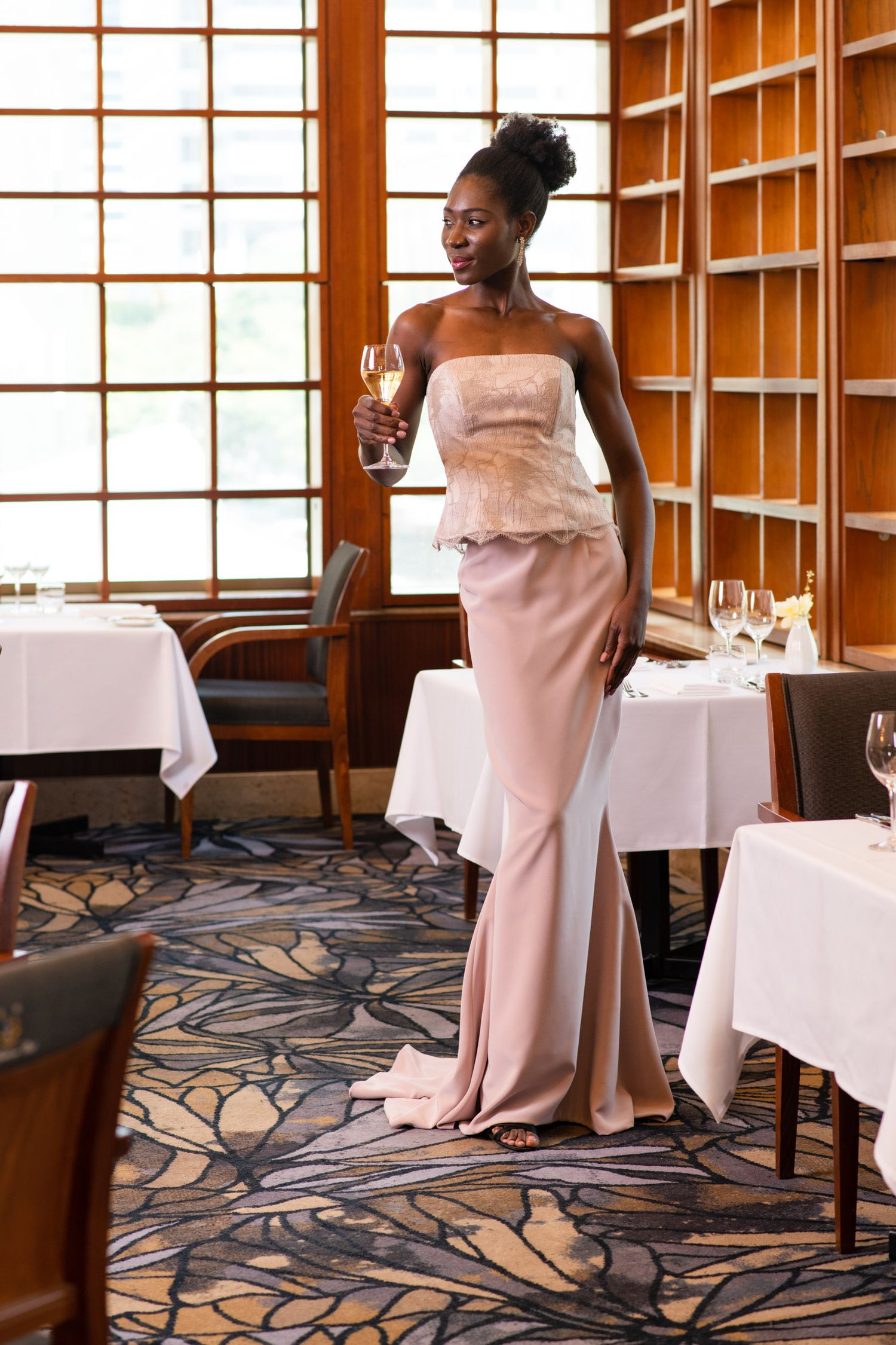 Tall woman in elegant pink gown holding a wine glass in an upscale restaurant dining room.