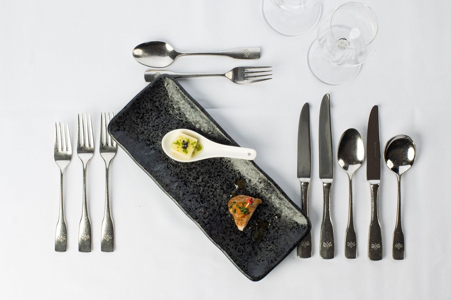 Fine dining table setting with black rectangular plate, gourmet bites, and polished silverware arrangement.