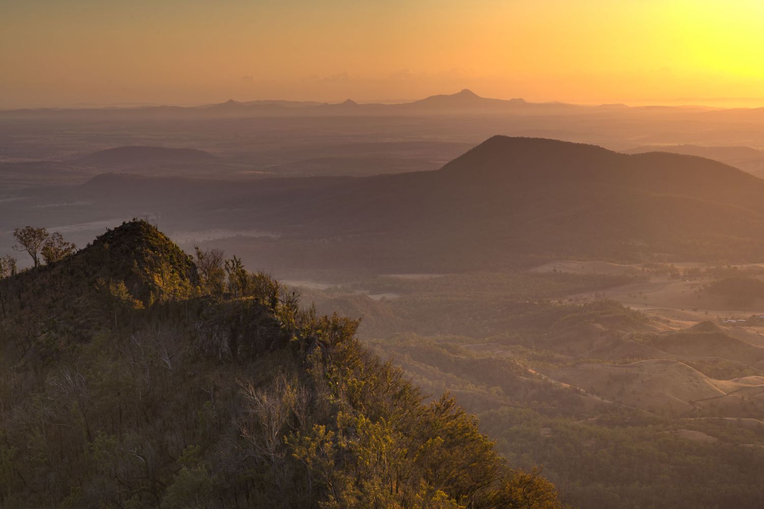Golden sunrise over misty mountain valleys with silhouetted peaks and lush vegetation in a scenic landscape.