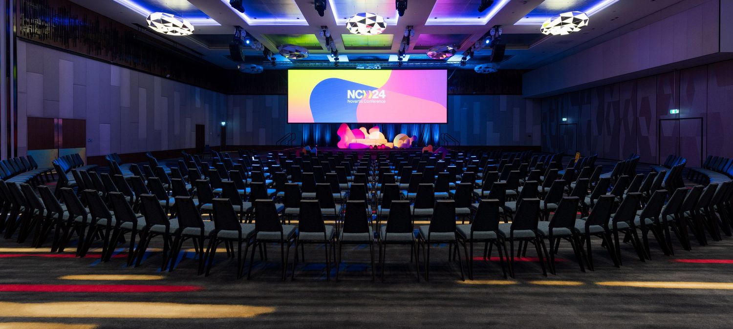 Large empty conference hall with rows of chairs facing a colorful illuminated stage screen at corporate event.
