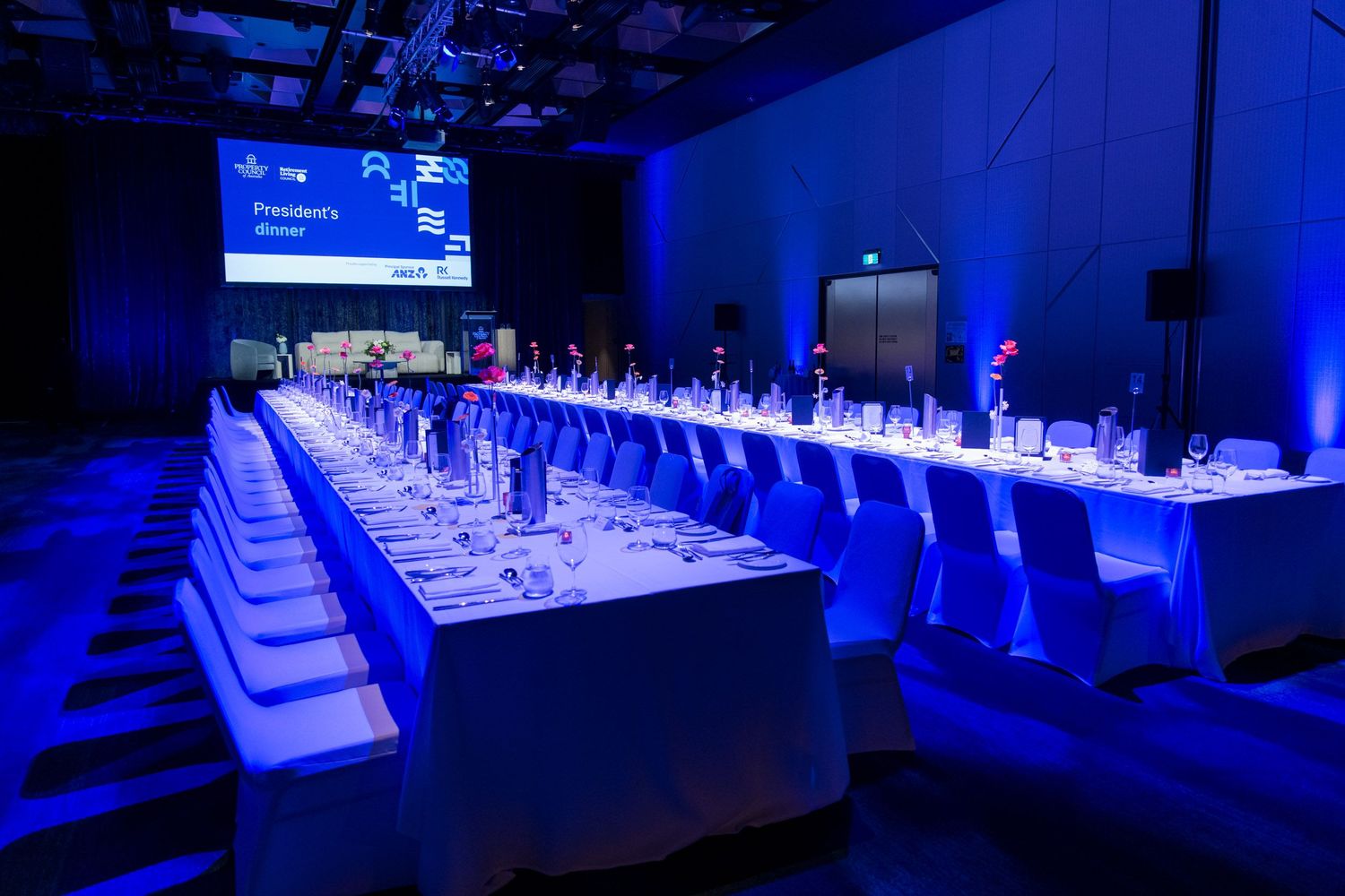 Long banquet table set for a Presidents Dinner with white linens and blue lighting in a dark event space.