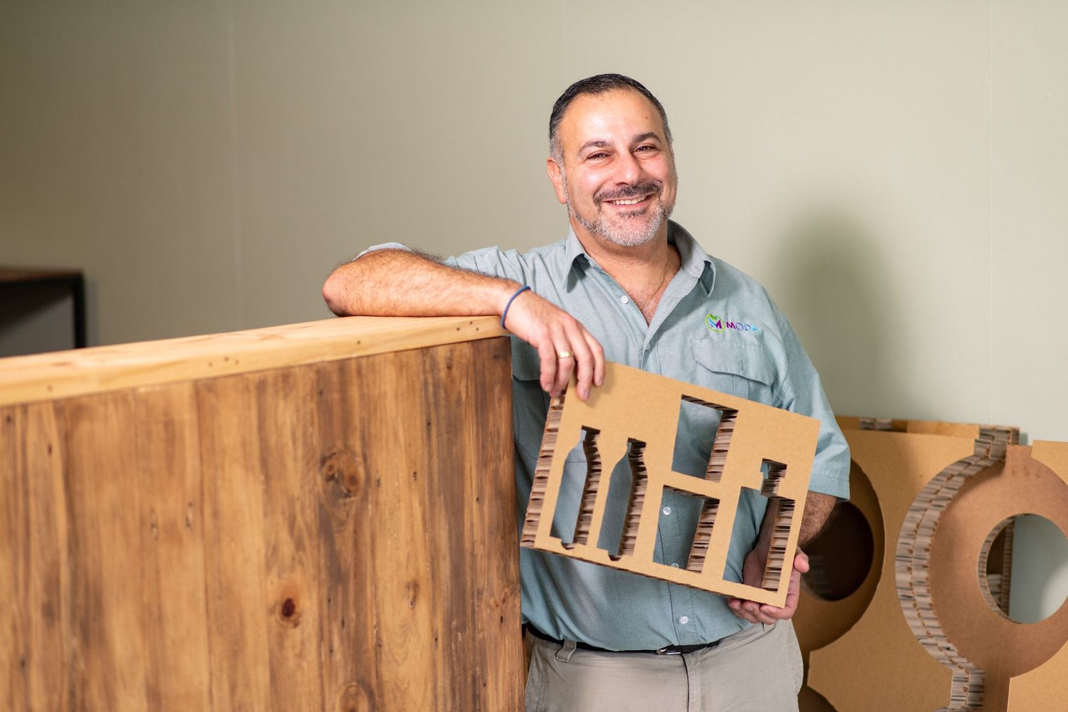 Smiling man holding a cardboard architectural model while leaning on a wooden counter.