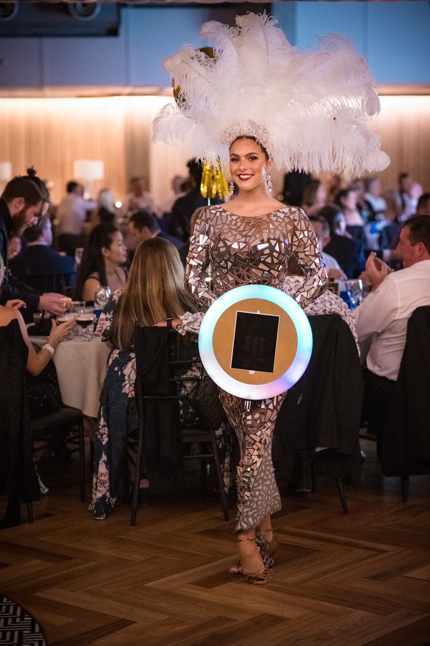 Glamorous performer in sequin gown and white feather headdress holding illuminated ring light at gala.
