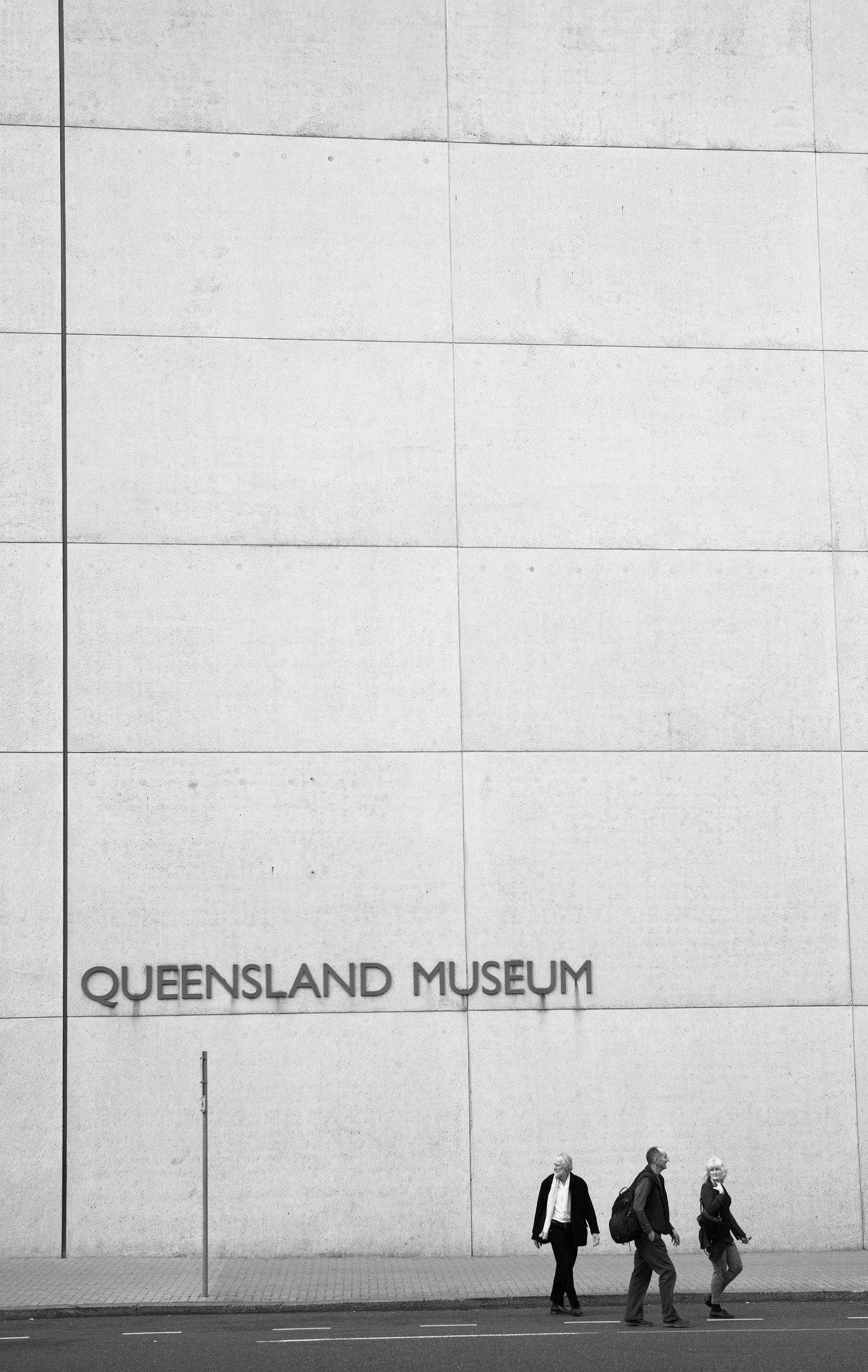 Three people walking past the large concrete wall of the Queensland Museum in black and white.