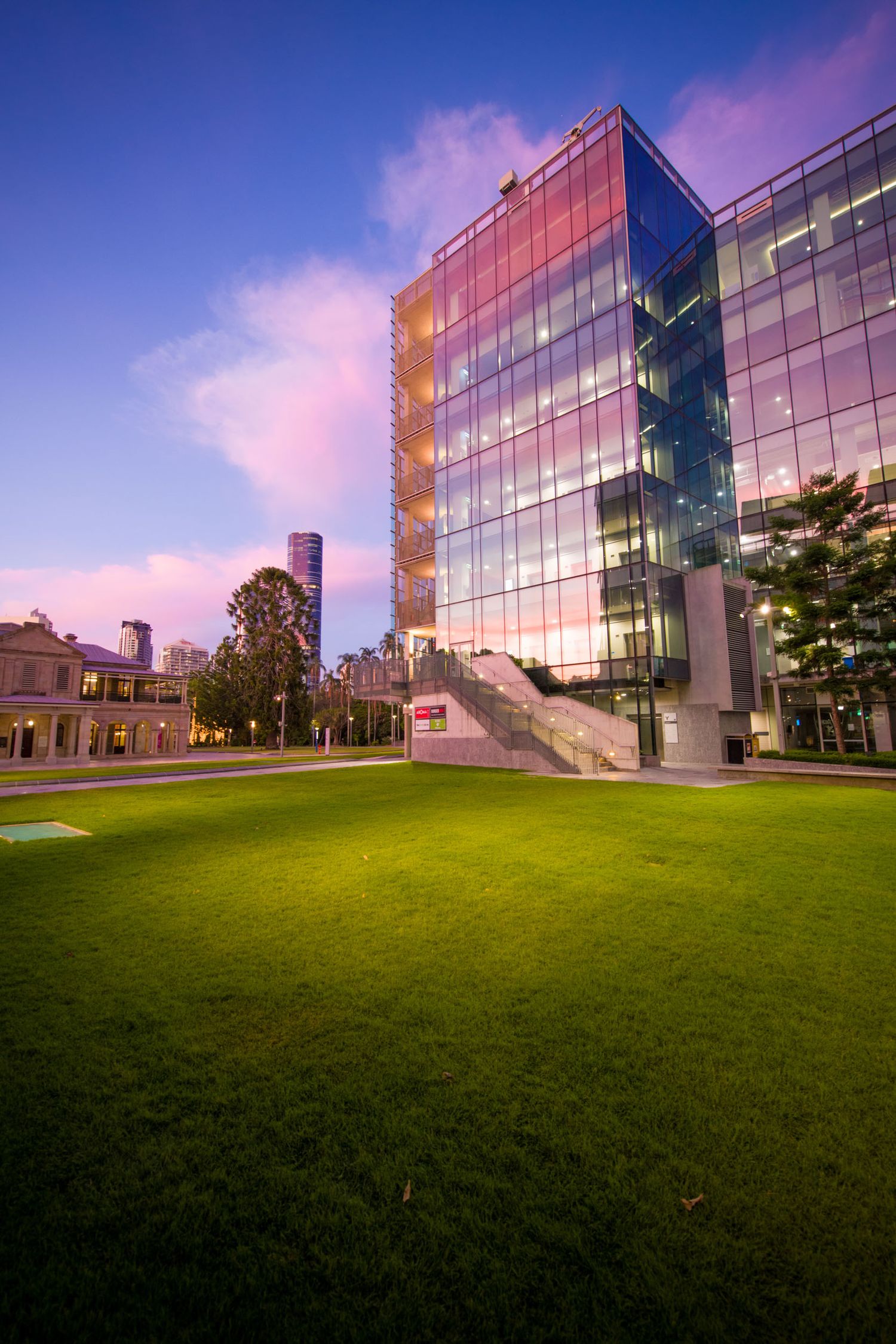 Modern glass office building reflecting colorful sunset sky beside a lush green lawn at dusk in an urban setting.