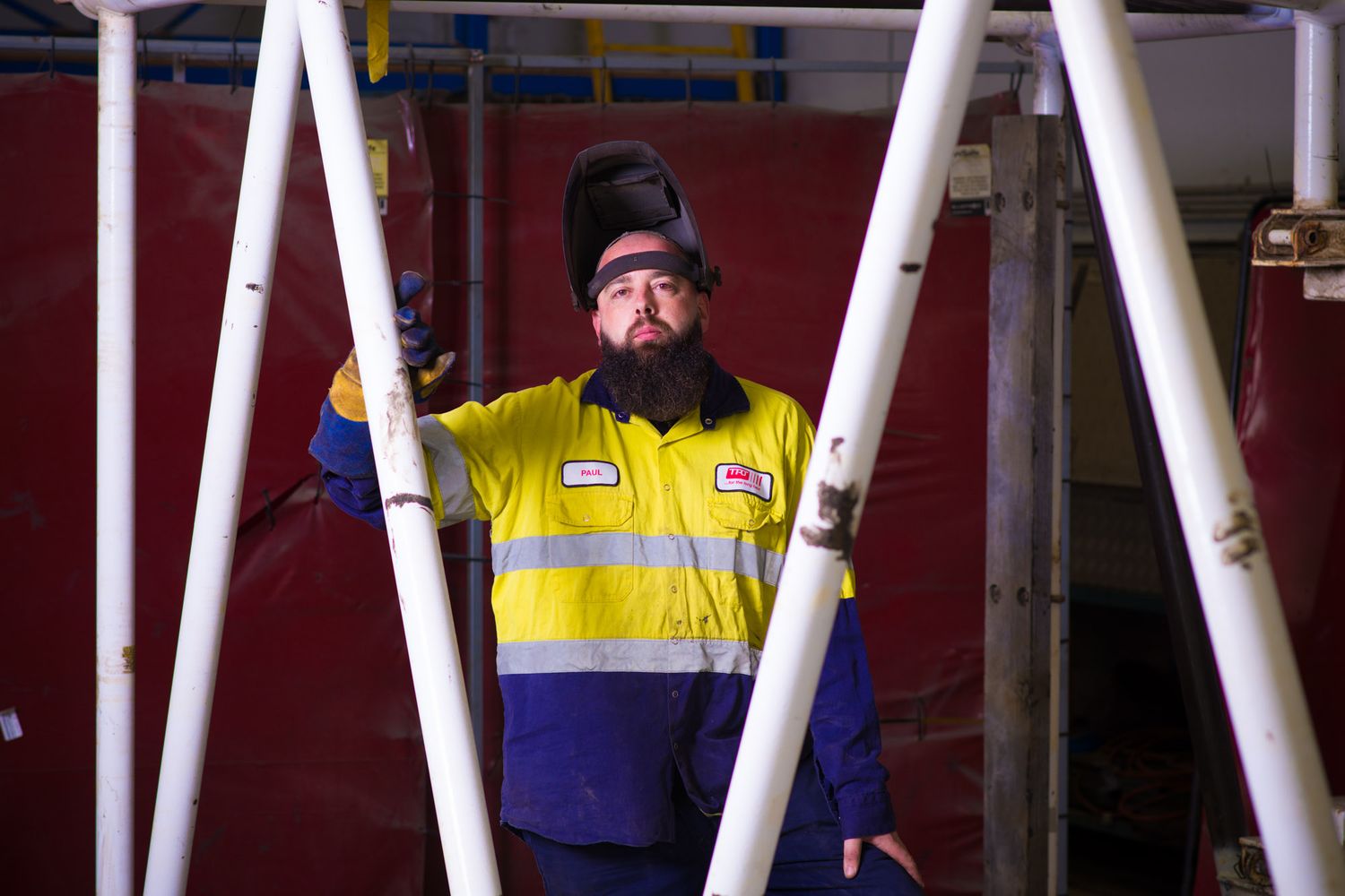 Bearded male worker in hi-vis uniform welding metal frames in an industrial workshop.
