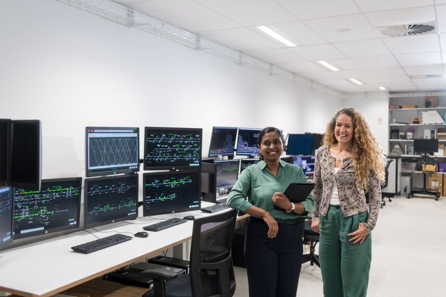Two professional women smile in modern control room surrounded by multiple computer monitors.