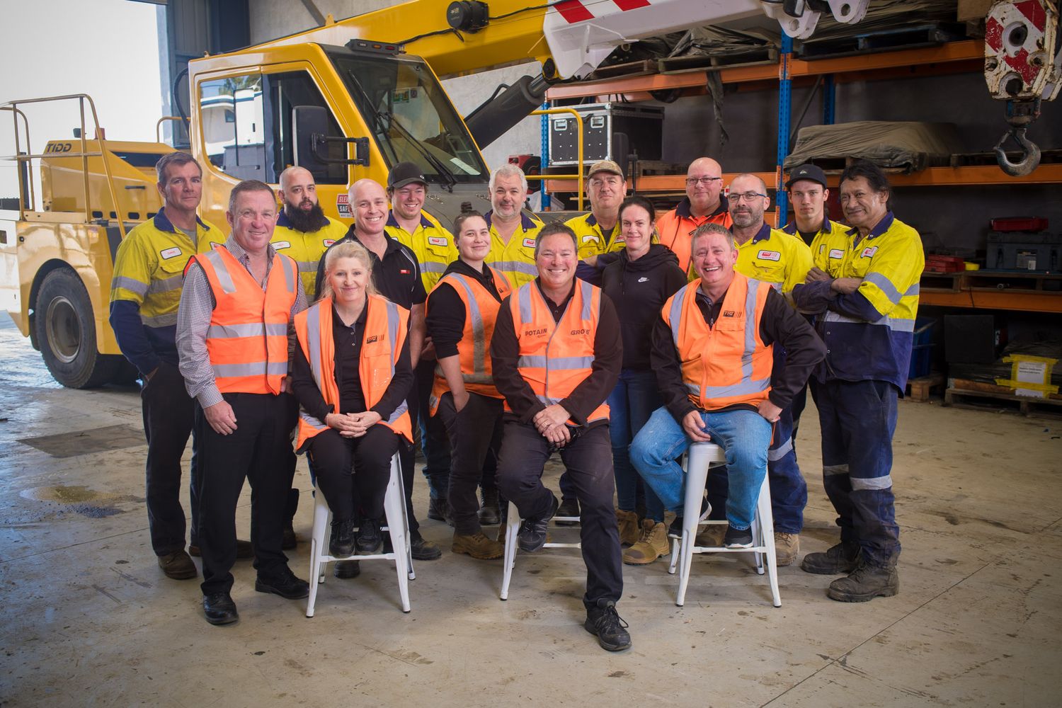 Large group of workers in hi-vis vests posing for team photo in industrial warehouse with yellow crane truck.