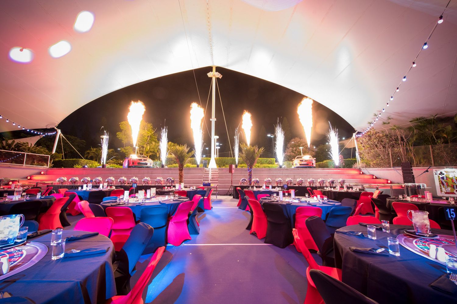 An elegant gala dinner tent setup with round tables, red accents, and dramatic pyrotechnic effects.