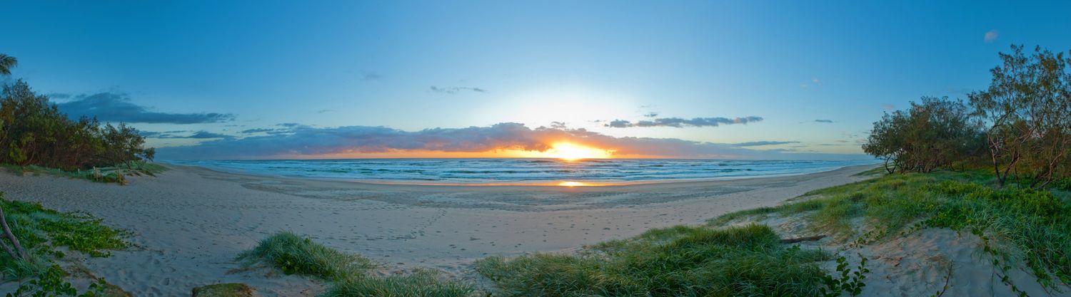 Panoramic view of peaceful sandy beach at sunset with golden sky reflecting on calm ocean water.