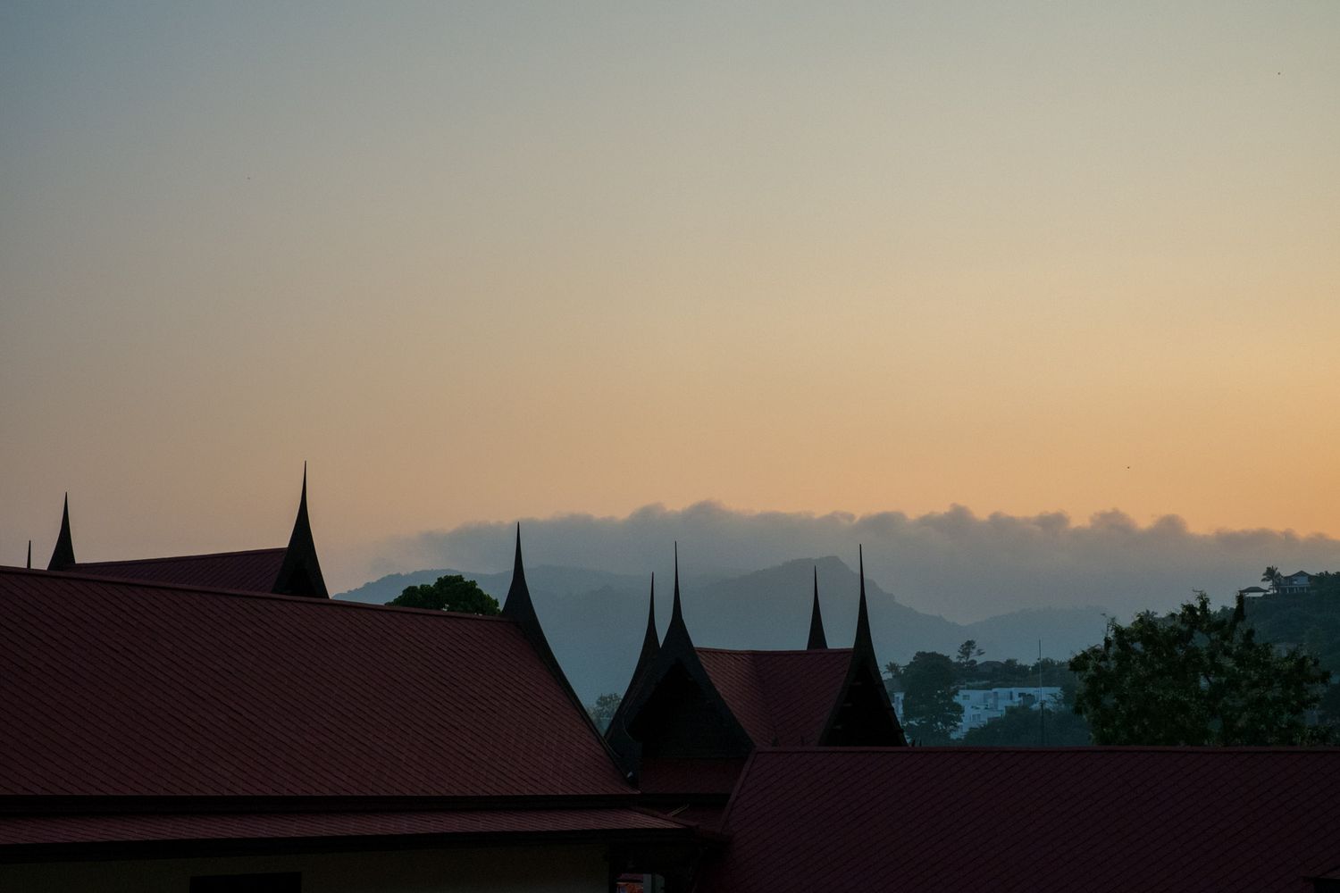 Silhouette of traditional Thai temple rooftops against a golden sunset sky with misty mountains in background.