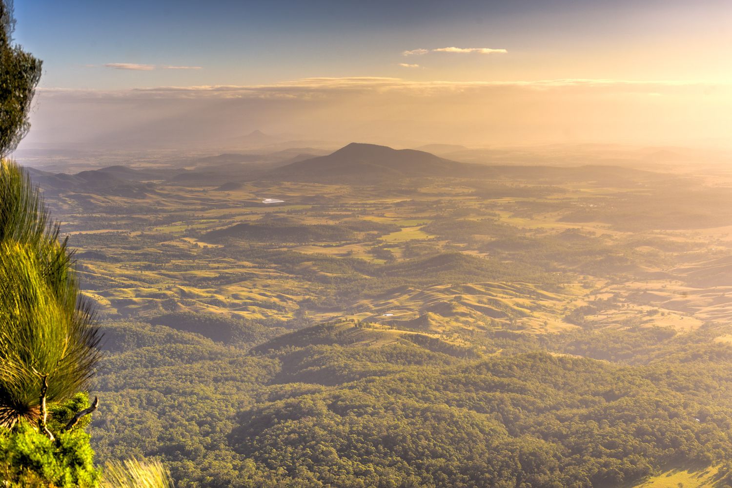 Golden hour panoramic view from a mountain summit overlooking vast green valleys and distant hills.