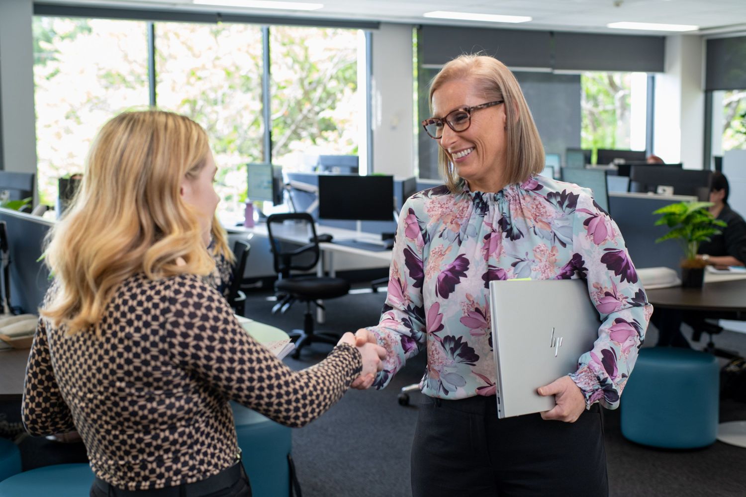 Two professional women shaking hands in a bright modern open-plan office, one holding a laptop and documents.