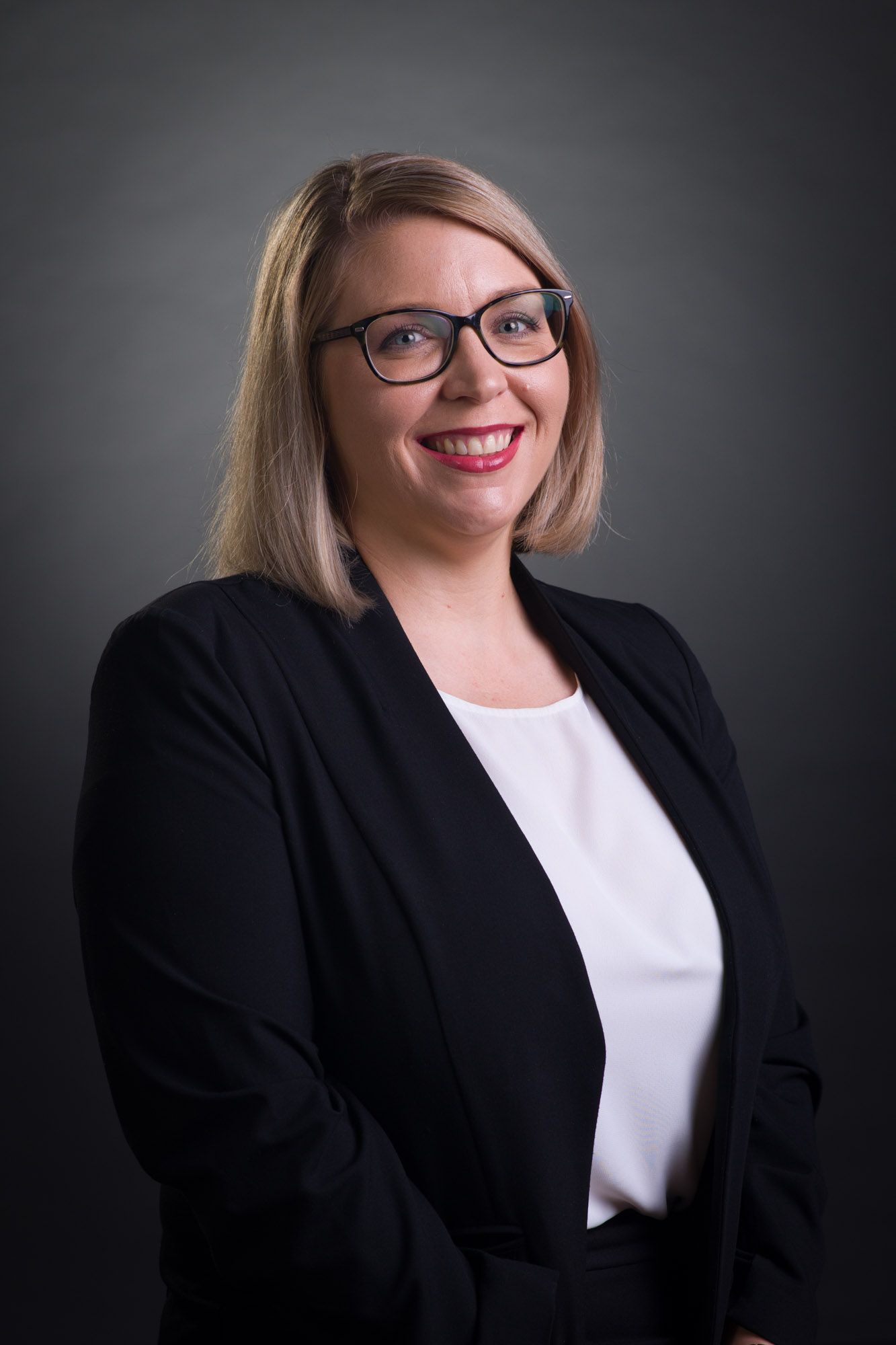 Smiling blonde woman in black blazer and glasses posing against a dark grey background.