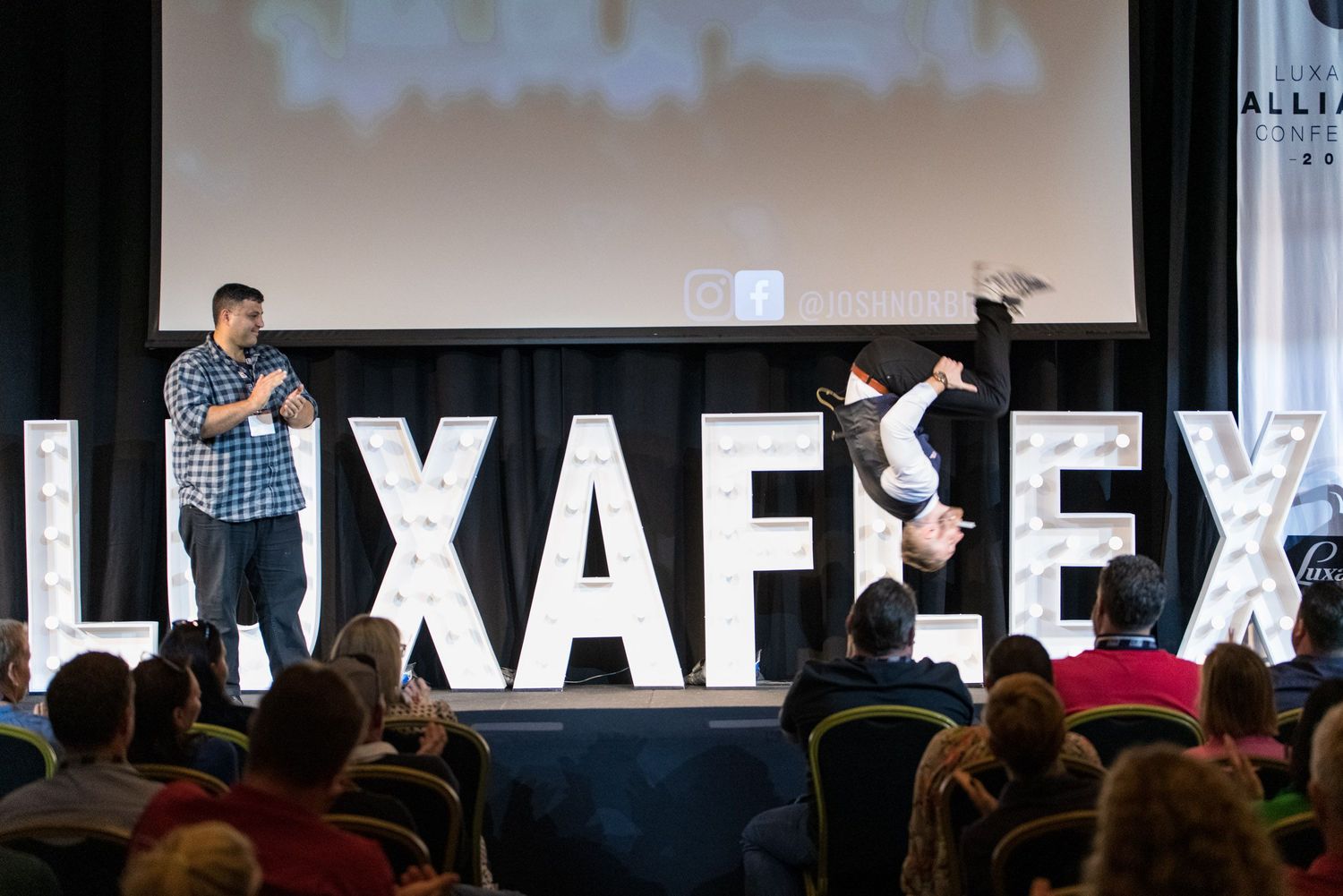 Two performers on stage in front of illuminated LUXAFLEX letters at a corporate conference event.