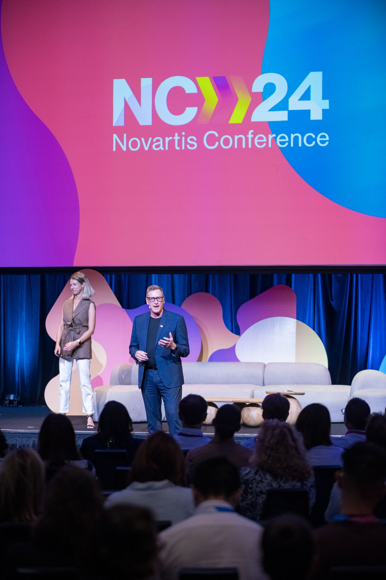 Two presenters stand on stage beneath the NC24 Novartis Conference logo at a corporate event.