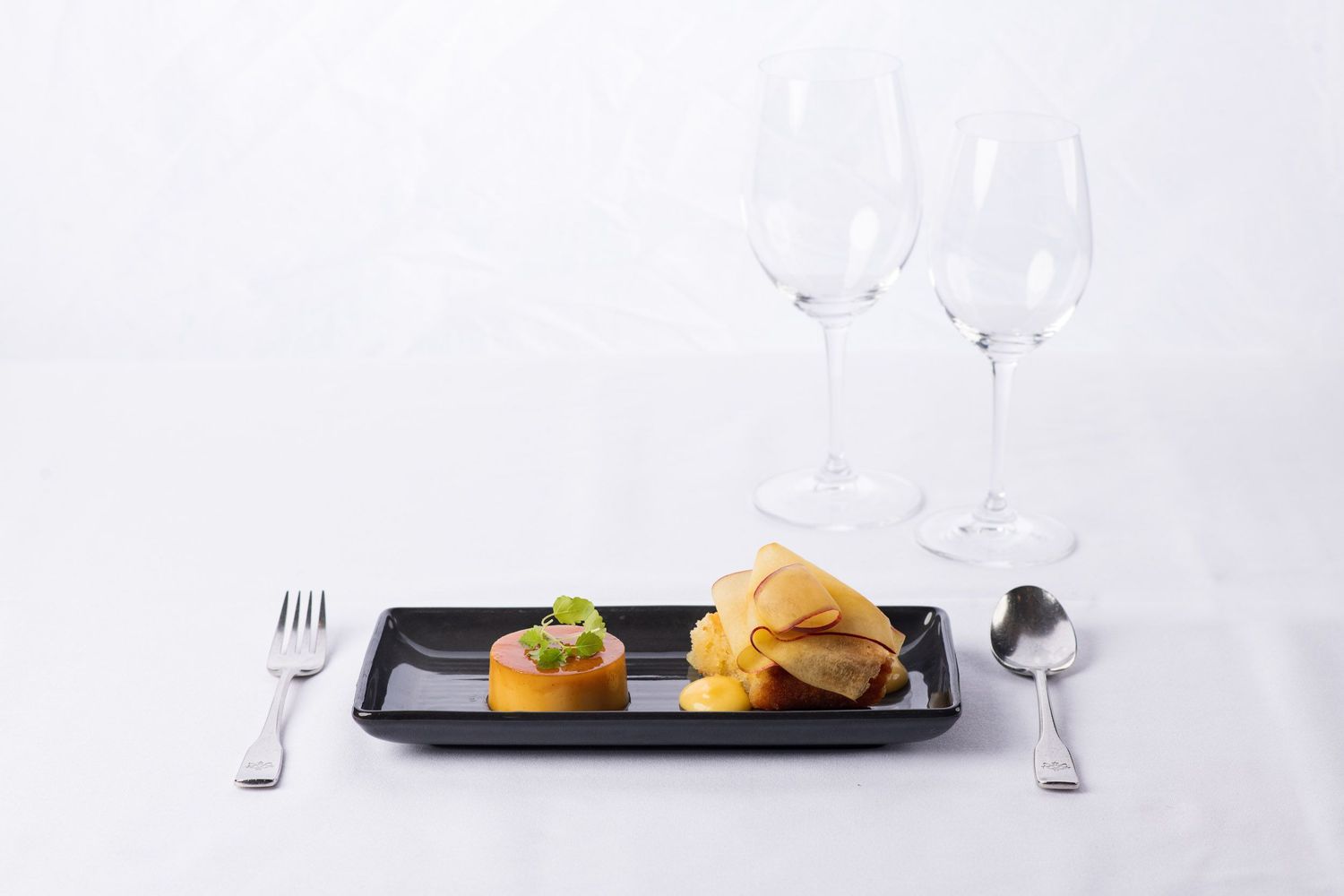Elegant restaurant appetizer course with two gourmet bites on black plate with crystal wine glasses.