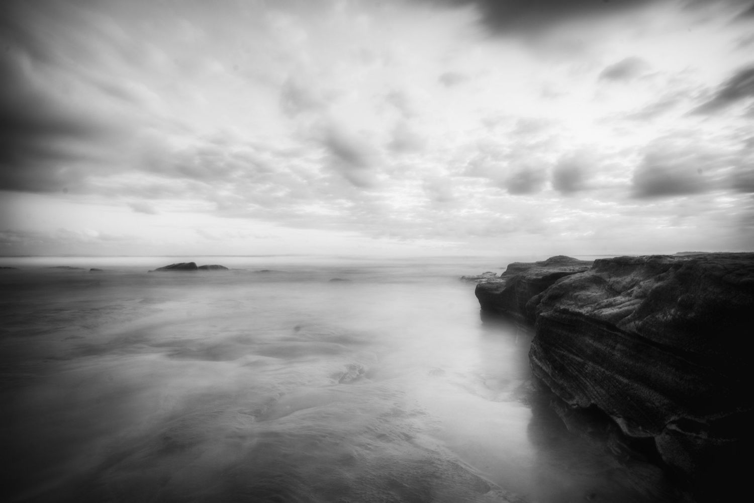 Black and white long exposure coastal seascape with rocky cliffs and dramatic cloudy sky.