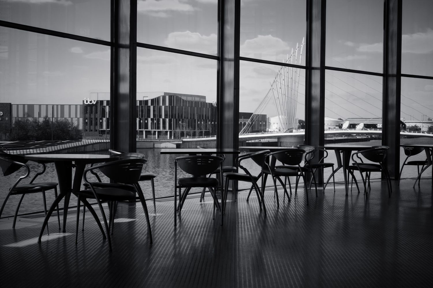 Black and white interior shot of empty cafe chairs and tables beside large floor-to-ceiling windows.