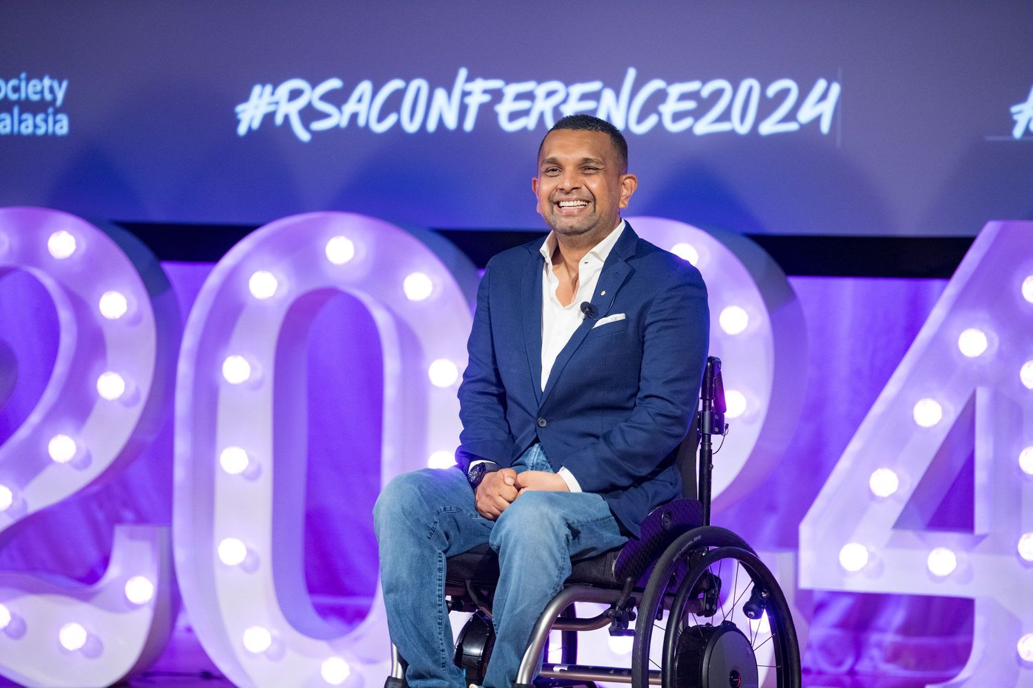 Smiling man in wheelchair poses before illuminated 2024 RSA Conference signage on stage.