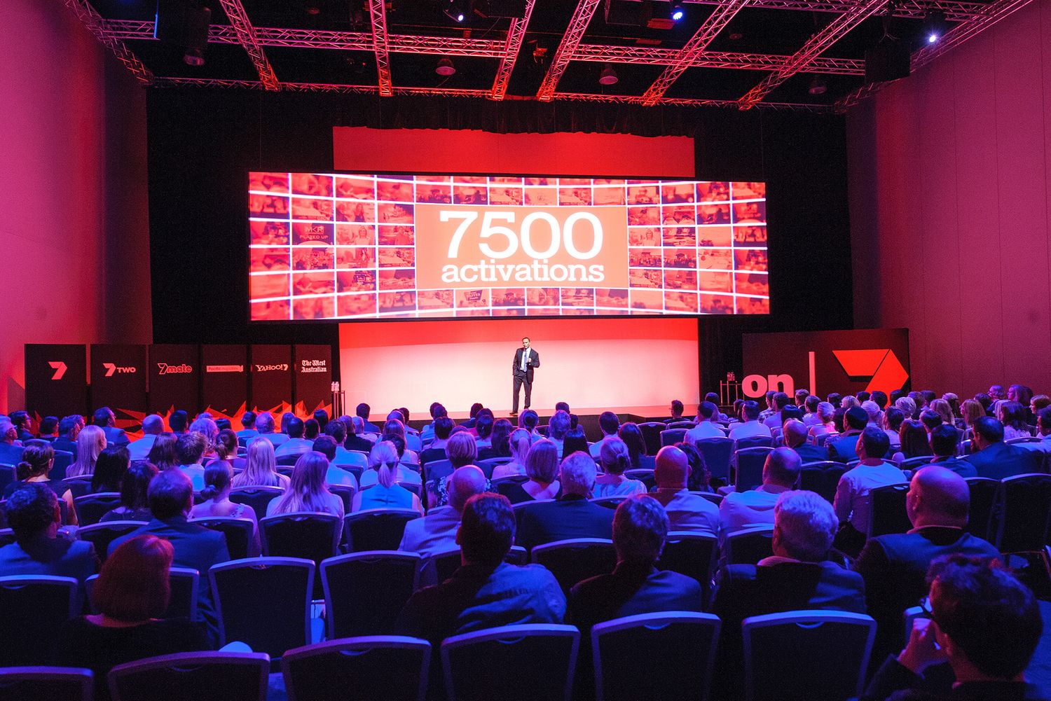 A large conference audience watches a presenter on stage with a screen displaying 7500 activations.