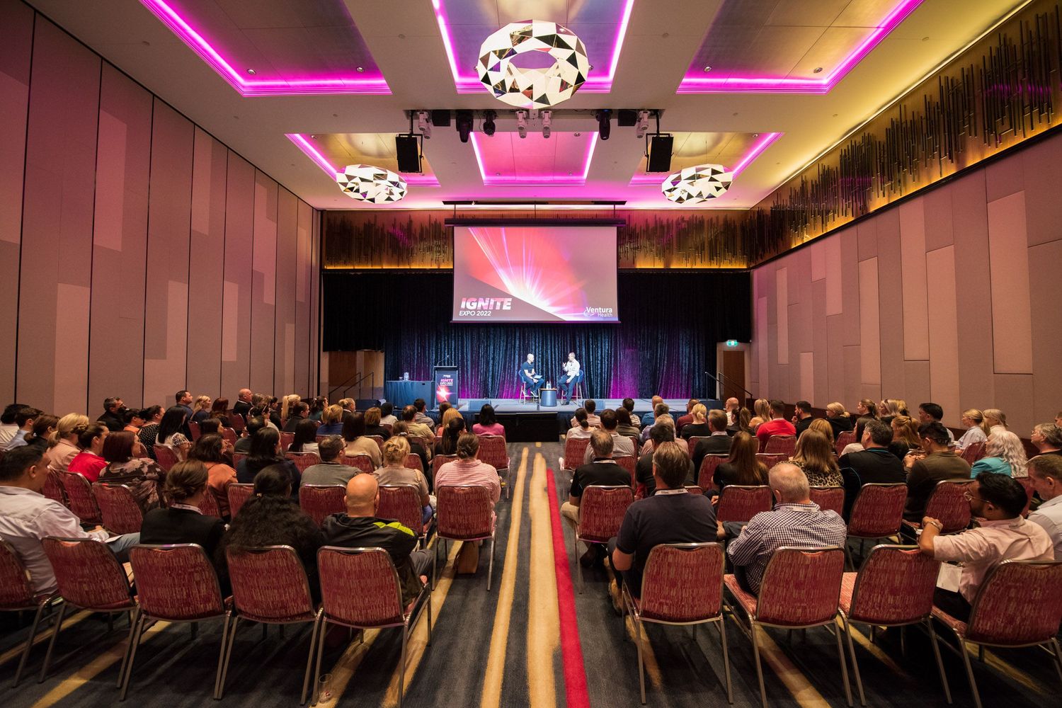 Large conference hall filled with attendees watching a presentation on a widescreen stage display.