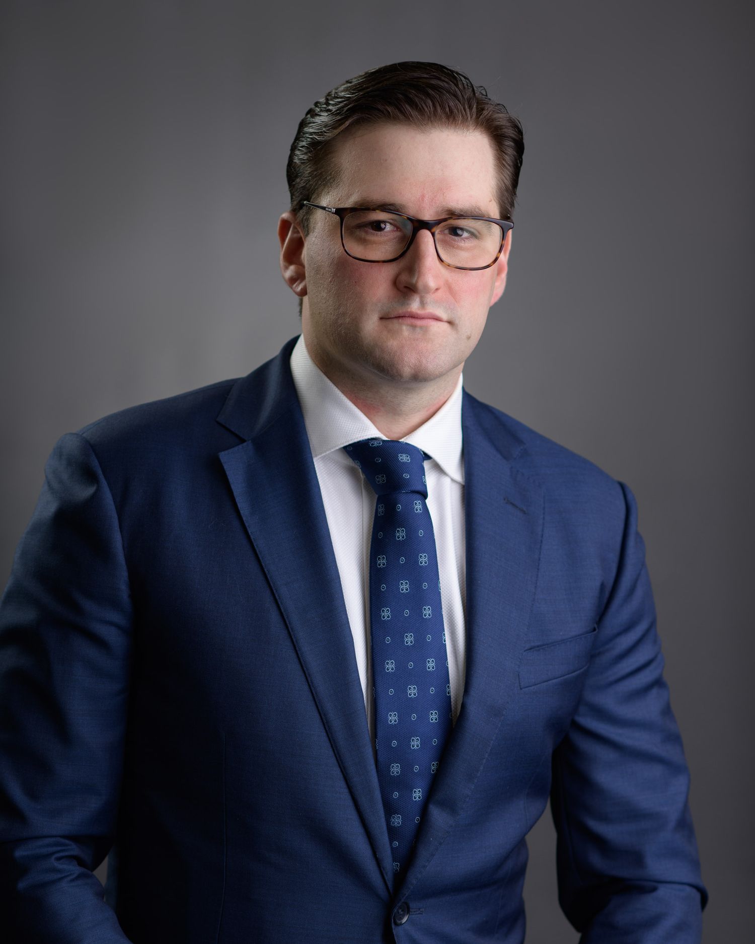 Professional man in blue suit and tie wearing glasses posing against grey studio background.