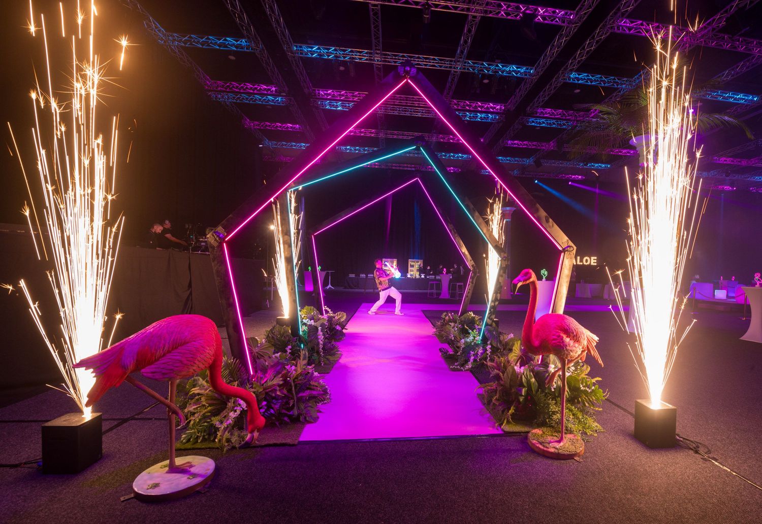 Dramatic gala entrance tunnel with neon pink arches, flamingo decor, and sparkling pyrotechnics on purple carpet.